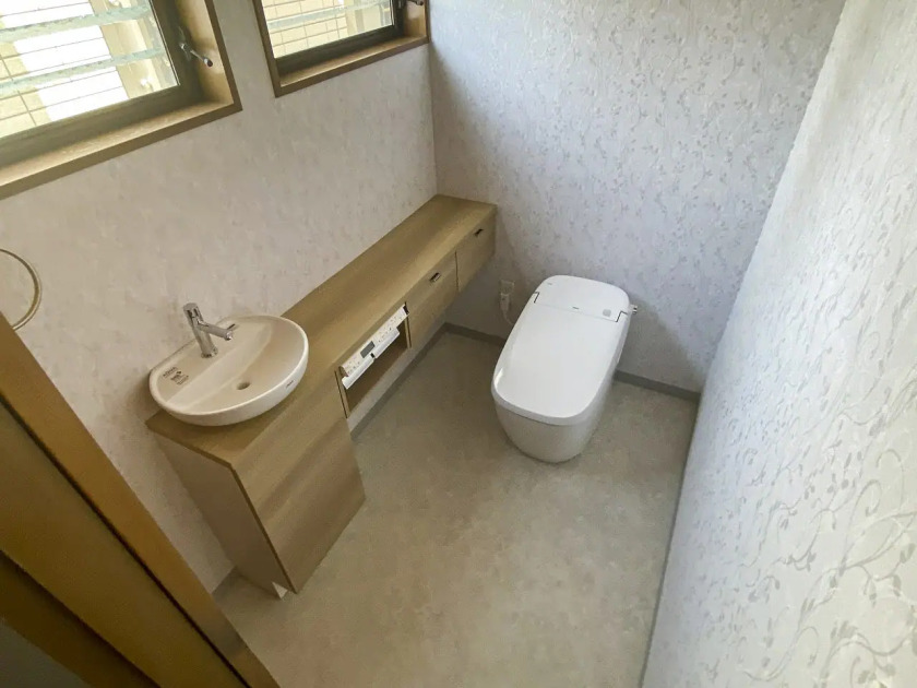 リフォーム後の施工事例。小便器を撤去し、LIXILのタンクレストイレ「サティスG」を設置。トイレ本体の位置を奥へ移動したことで、左側の壁面に手洗い器付きの木目調ロングカウンター収納を新設。壁紙は上品な花柄のクロスに貼り替えられ、広々とした機能的で清潔感のあるトイレ空間。