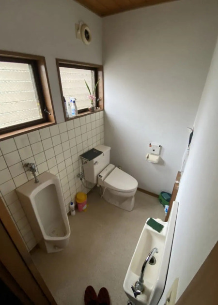 一戸建てのトイレリフォーム前の様子。同じ空間内に、男性用の小便器（左）と、手洗いタンク付きの洋式トイレ（奥）が並んで設置されている。壁面は白いタイル貼りで、窓際や棚には掃除用具などが置かれており、複数の設備が並ぶことでやや手狭な印象の内装状態。