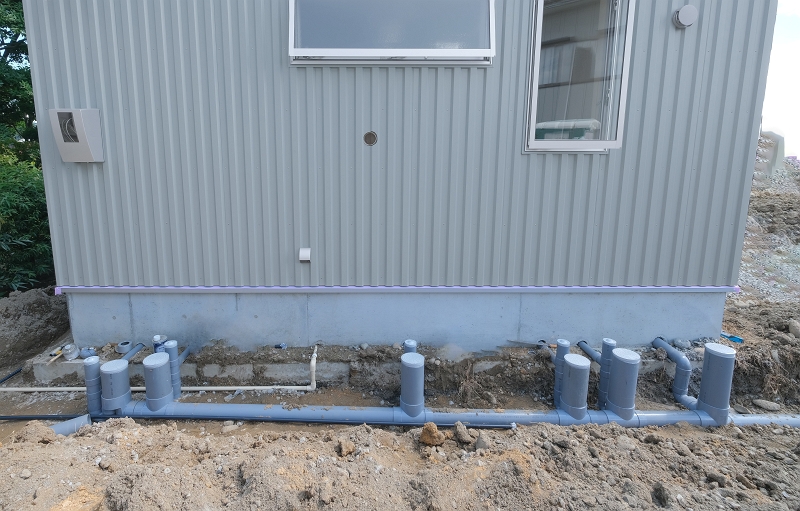 建物の基礎外周に敷設された屋外排水配管の施工現場。グレーの硬質塩化ビニール管が地面に配置され、等間隔に複数の排水桝（はいすいます）の立ち上がり管が設置されている。建物からの排水を公共下水道へスムーズに流すための、耐用年数が高くメンテナンス性に優れた屋外配管設備の様子。