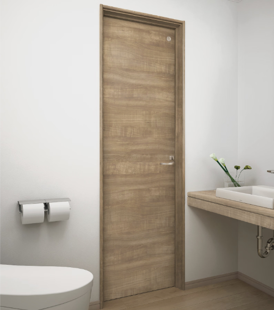 DAIKENのトイレドア「ieria（イエリア）」の設置事例。銘木の質感をリアルに再現したライトブラウンの木目調ドアが、白を基調とした清潔感のあるトイレ空間に設置されている。横には木製カウンターの手洗器とシルバーのペーパーホルダーが配置され、マットで上質な素材感が落ち着いたインテリアを演出している様子。