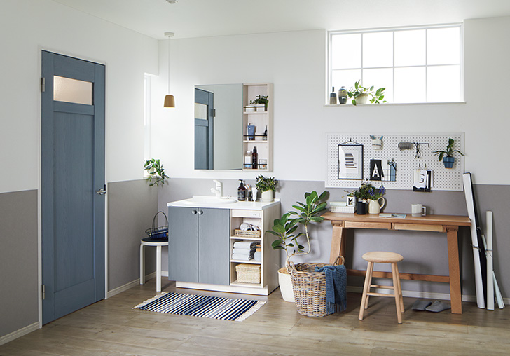 Panasonicのドア「ベリティス」を中心とした、洗面・ワークスペースのコーディネート例。ネイビーブルーの木目調ドア（採光窓付き）と、同色の扉を採用した洗面化粧台が統一感のある空間を演出。壁面は白とグレーのツートンカラーで、木製のデスクや観葉植物、有孔ボードが並ぶ、機能的で清潔感のあるインテリア。